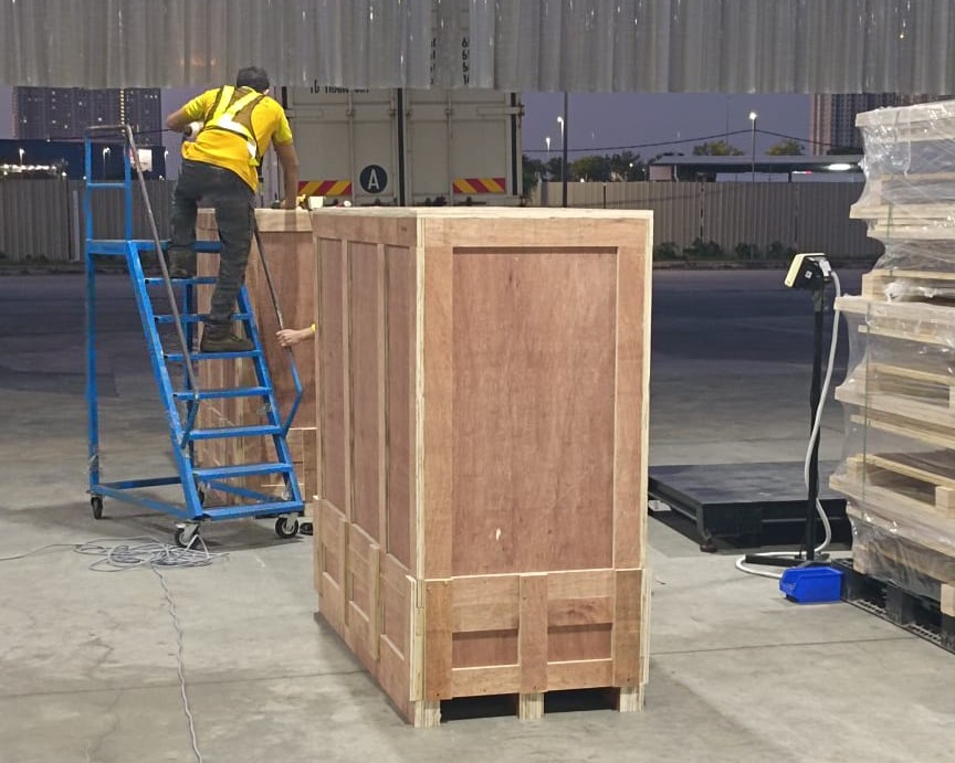 M&C crew loading a tall plywood export crate beside a shipping container