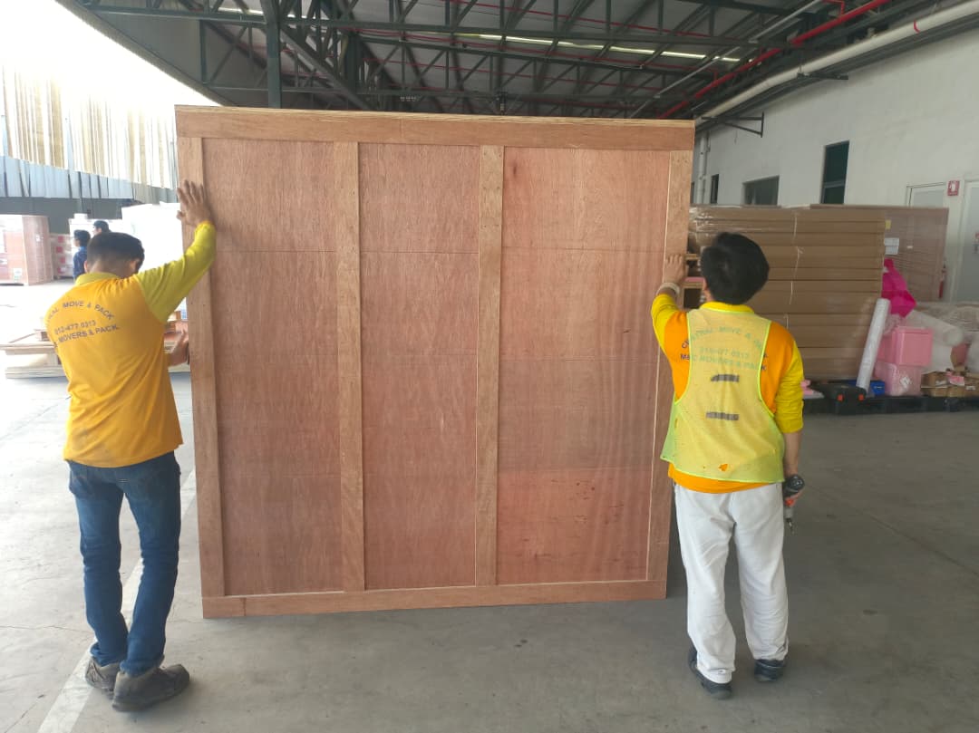 Two M&C crew in branded uniform positioning a large custom wooden plywood crate in a Penang warehouse