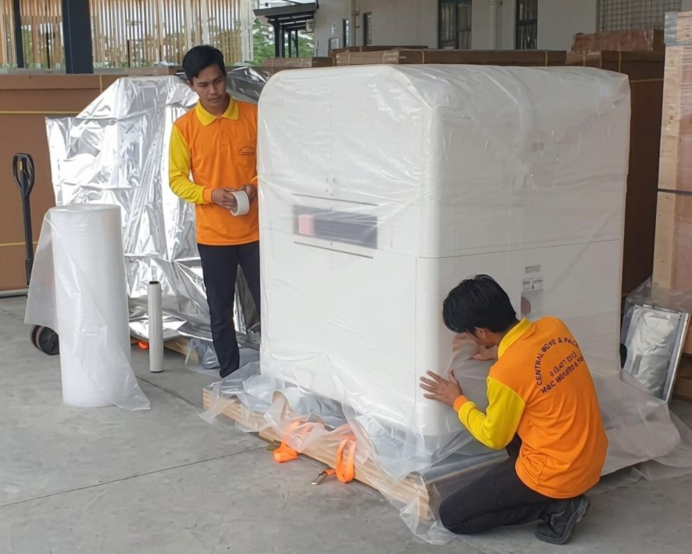 An M&C worker carefully wrapping a large machine in protective foam and film before transport